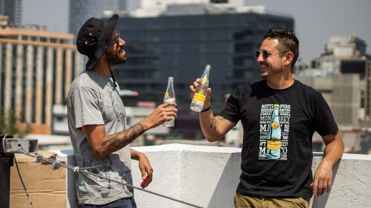 Dos hombres están de pie en una azotea en una ciudad, sosteniendo botellas de Topo Chico. Un hombre lleva un sombrero y una camisa gris claro, mientras que el otro lleva gafas de sol y una camiseta negra con un diseño de Topo Chico. Edificios son visibles en el fondo.