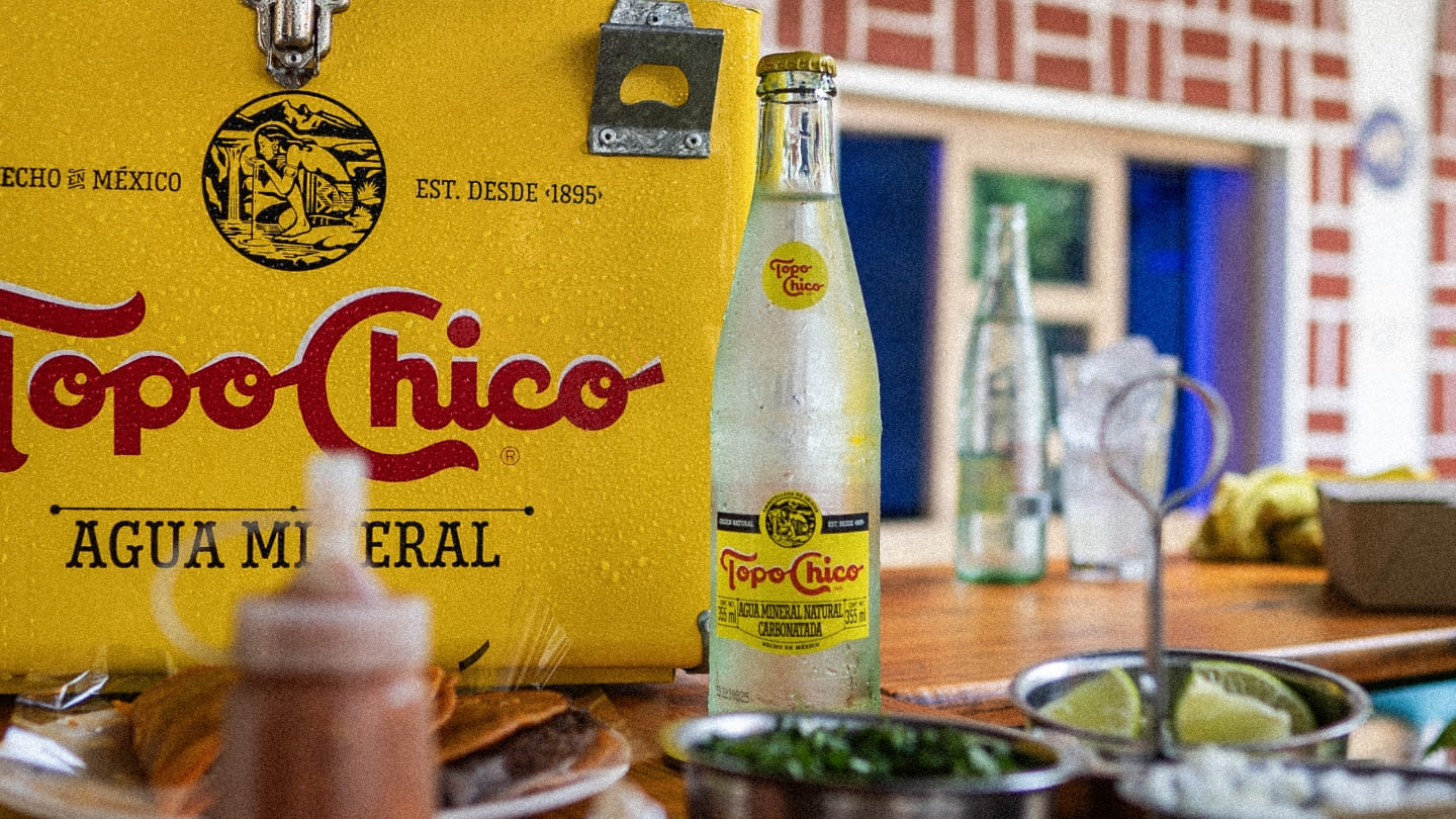 Una foto de primer plano de una botella helada de Topo Chico frente a una hielera amarilla de Topo Chico, con tacos y condimentos en primer plano, evocando una escena mexicana refrescante y sabrosa.