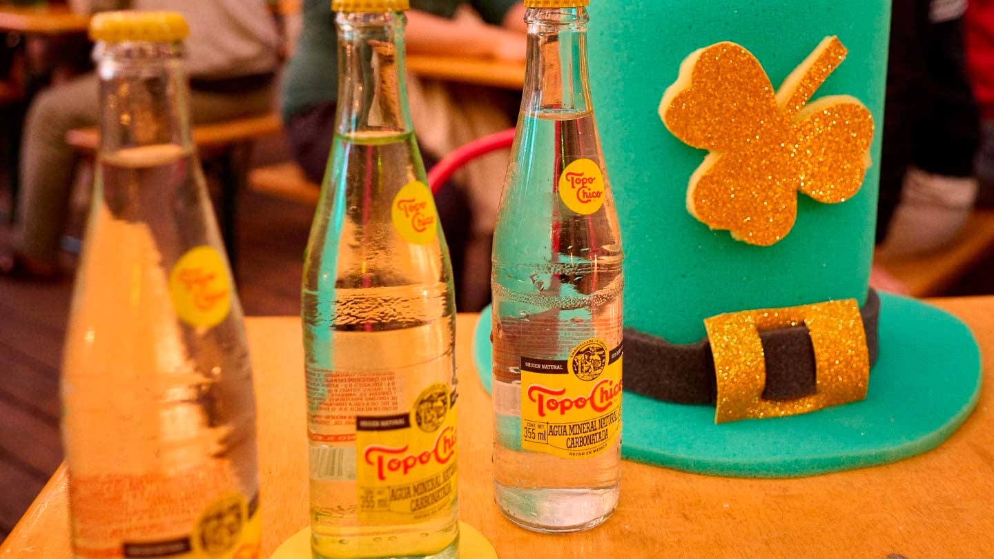  Tres botellas de Topo Chico se sientan junto a un sombrero verde de San Patricio con un trébol dorado, creando una escena festiva y refrescante.