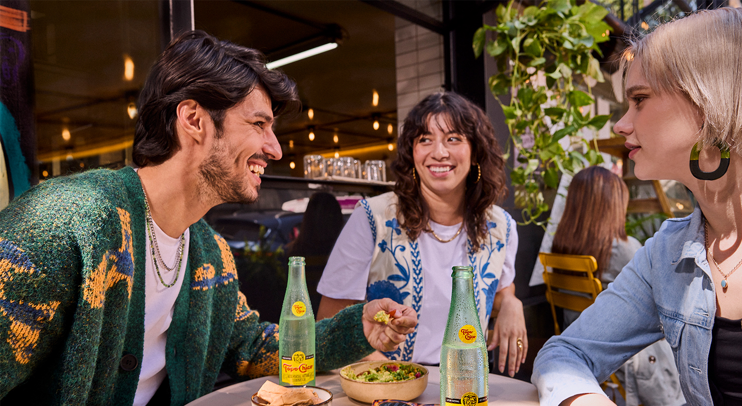 Tres amigos disfrutan de comida y Topo Chico en un café al aire libre.