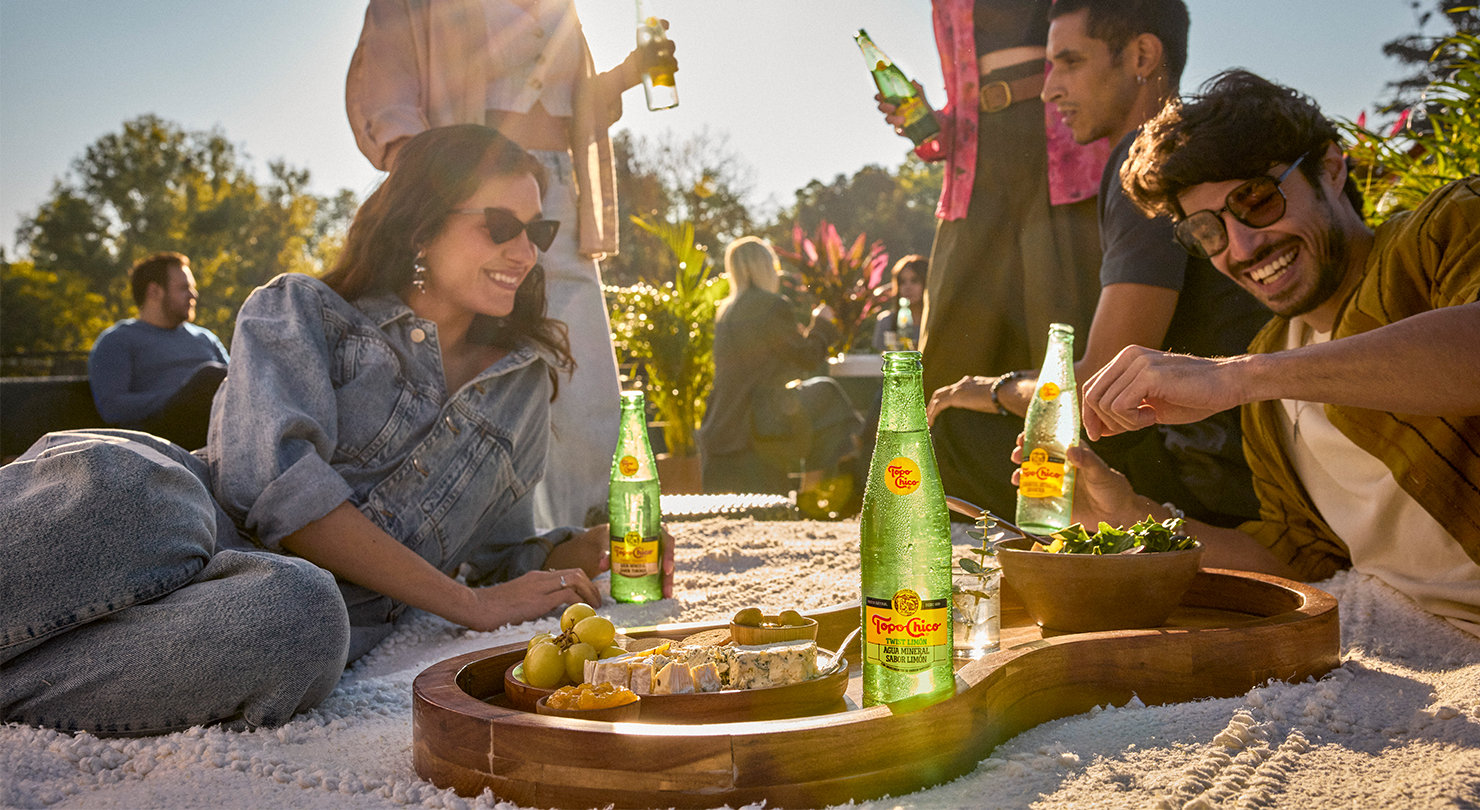 Un grupo de amigos disfruta de un picnic soleado con Topo Chico, una variedad de quesos, uvas, ensalada y otros bocadillos dispuestos en una bandeja de madera.