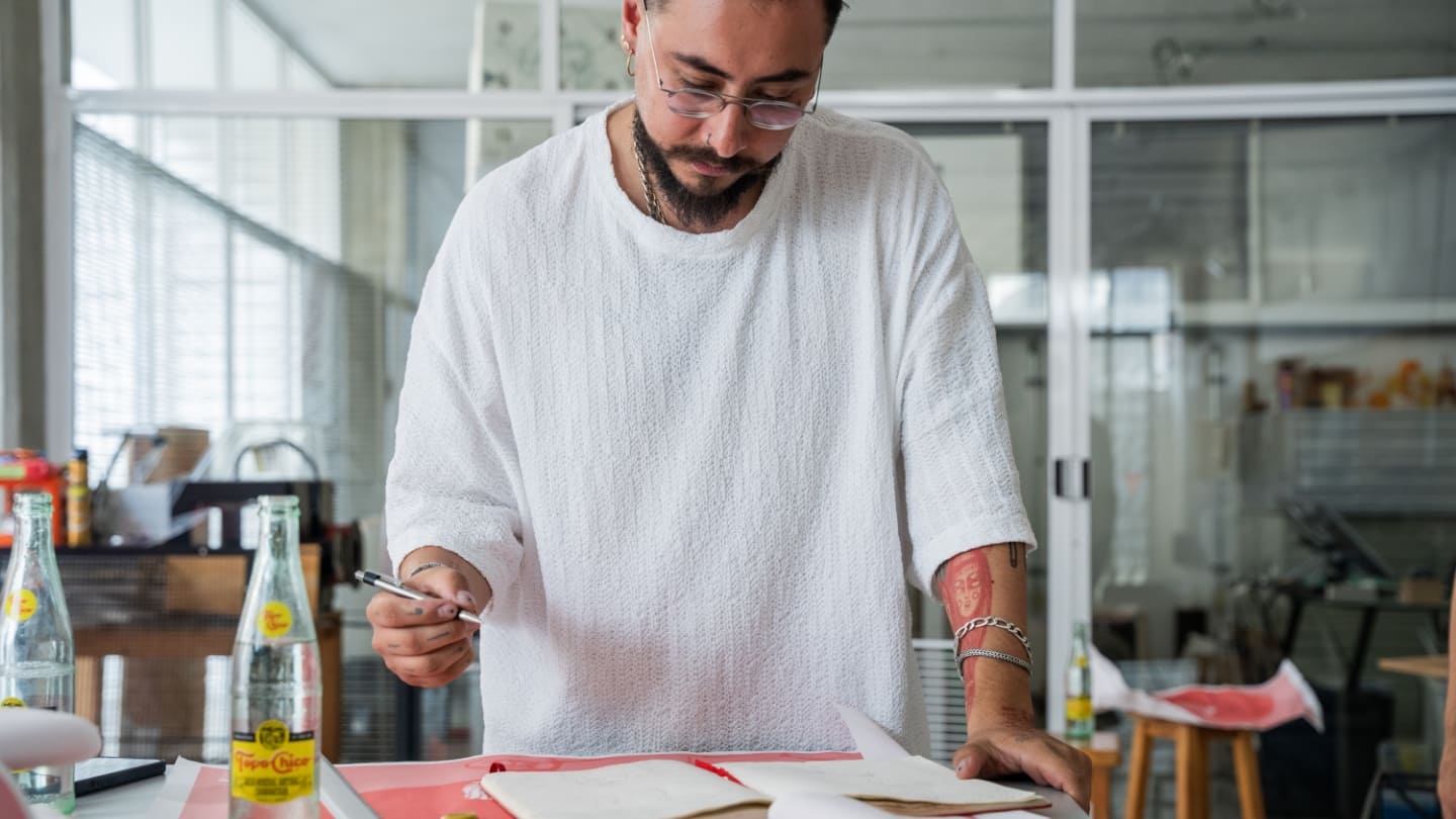 Diseñador parado en frente de una mesa, escribiendo en un cuaderno y con una botella de vidrio Topo Chico