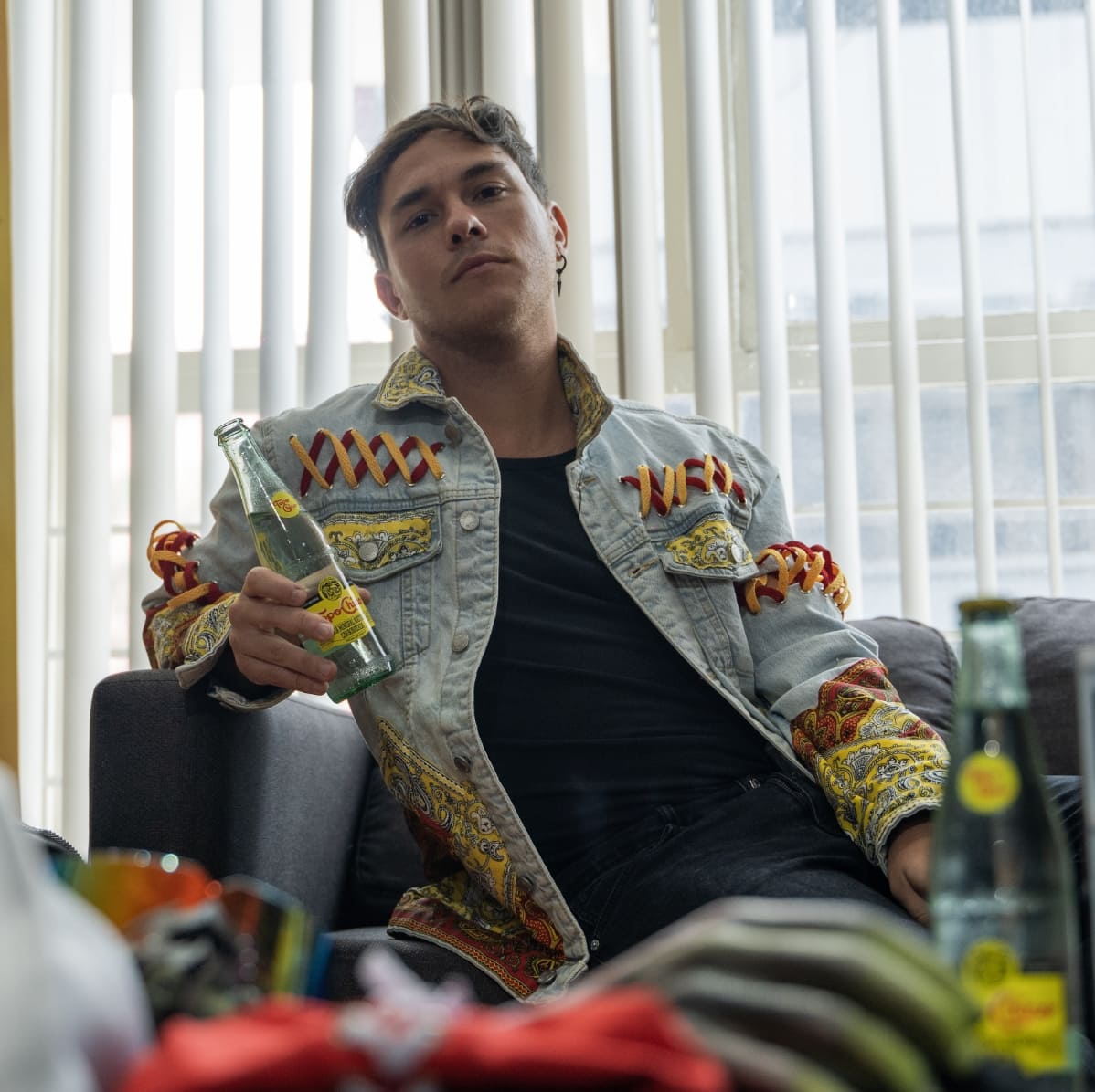 Persona sentada en un sofá, usando una Jean Jacket con cintas de color rojo y amarillo, sosteniendo una botella de vidrio Topo Chico