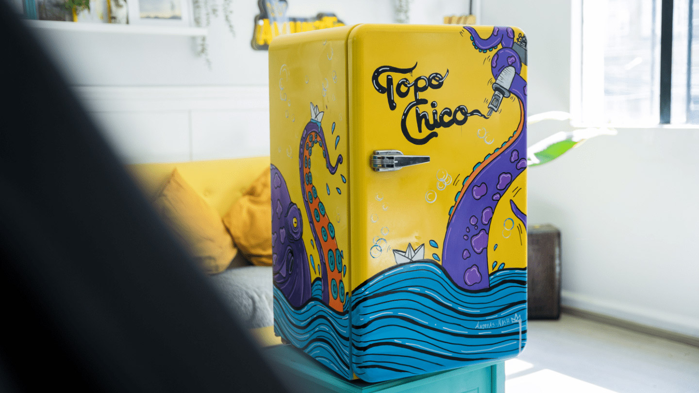 Heladera con logo Topo Chico, pintada de amarillo con un mar, un barquito de papel y un pulpo violeta.