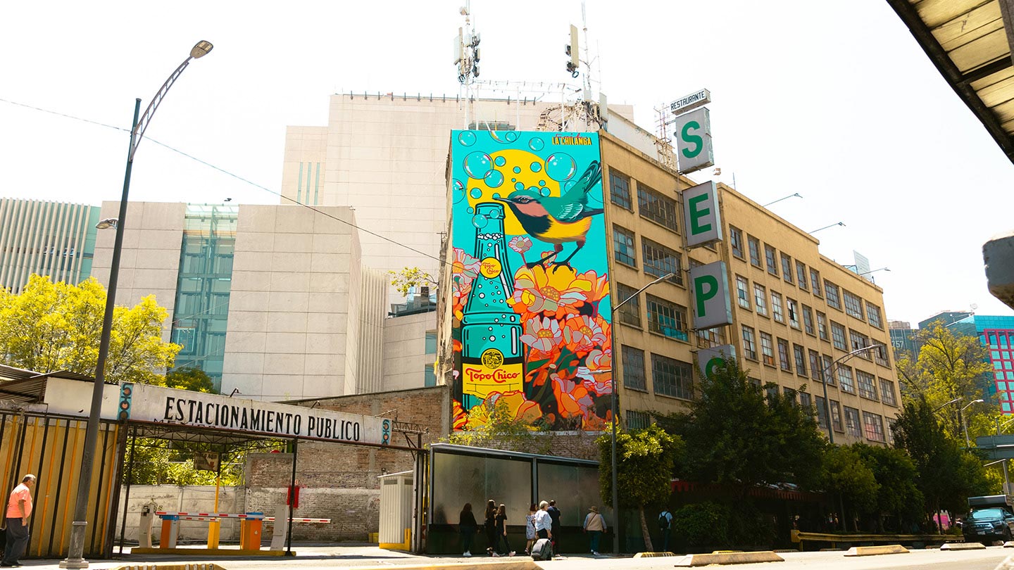 Pintura wn el lateral de un edificio, es una botella Topo Chico con un pájaro en colores verde agua y amarillo.