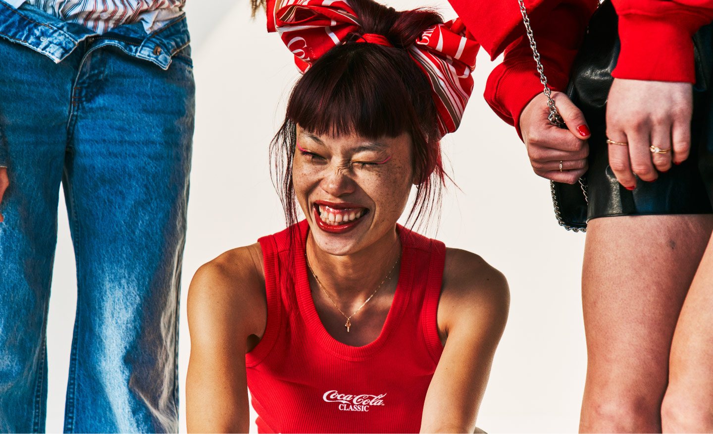 Persona sonriendo con top rojo y maquillaje colorido, sentada entre otras personas durante una sesión fotográfica de Coca-Cola.