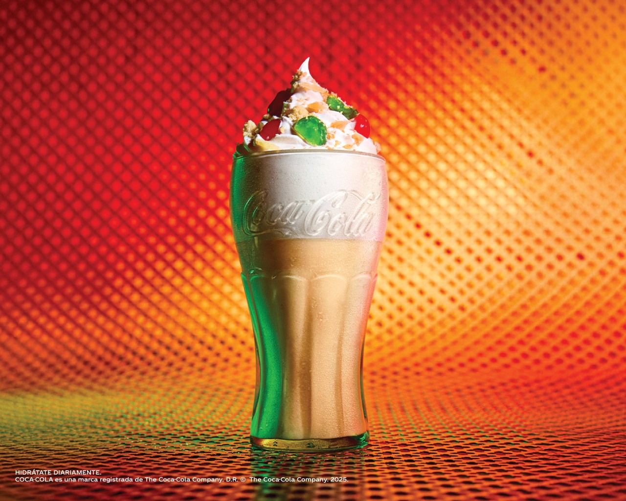 Bebida especial de Coca-Cola con crema batida y decoraciones de colores, presentada en vaso de vidrio sobre base decorativa.​