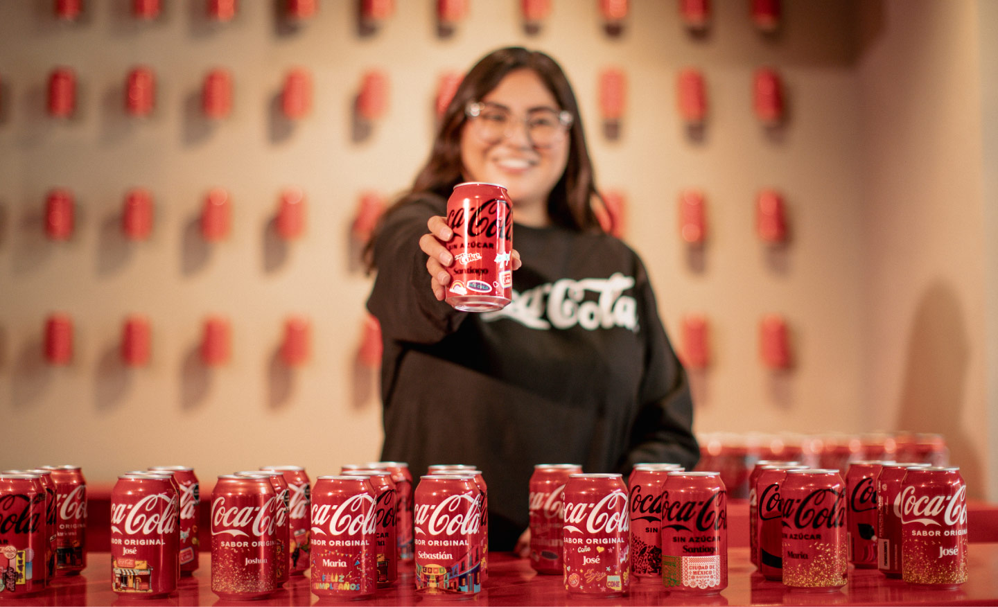 Persona sonriendo ofrece una lata de Coca-Cola al frente, con varias latas de la marca alineadas sobre una mesa.​