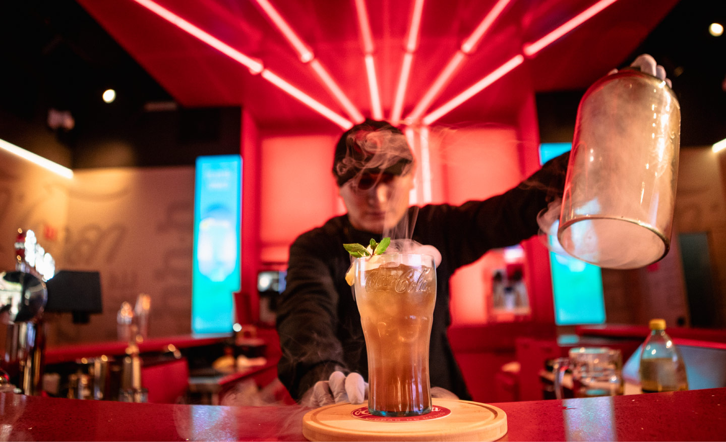 Bartender prepara una bebida exclusiva en House of Coca-Cola, con vaso alto, humo decorativo y barra iluminada en tonos rojos.​
