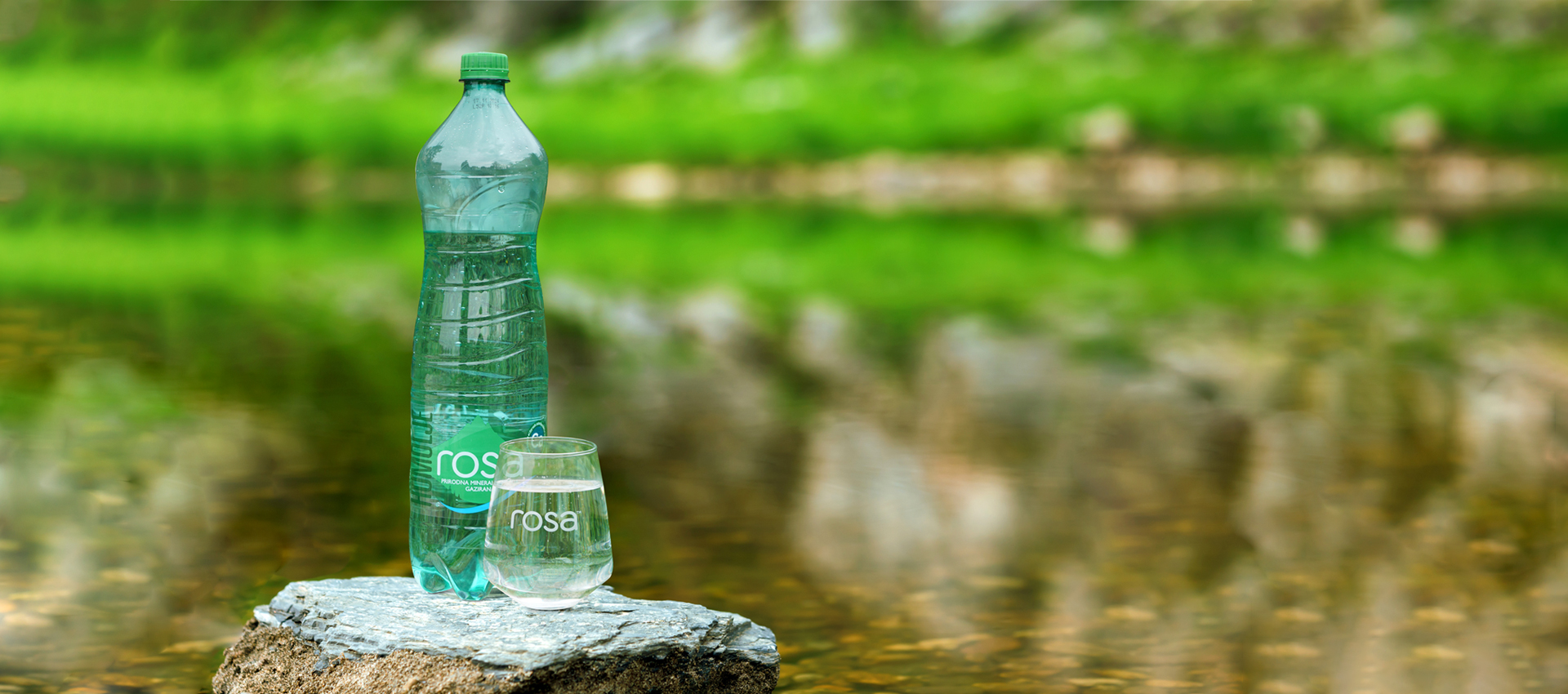 Flaša Rosa vode i čaša sa vodom postavljene na kamen pored bistre reke, sa zelenilom u pozadini