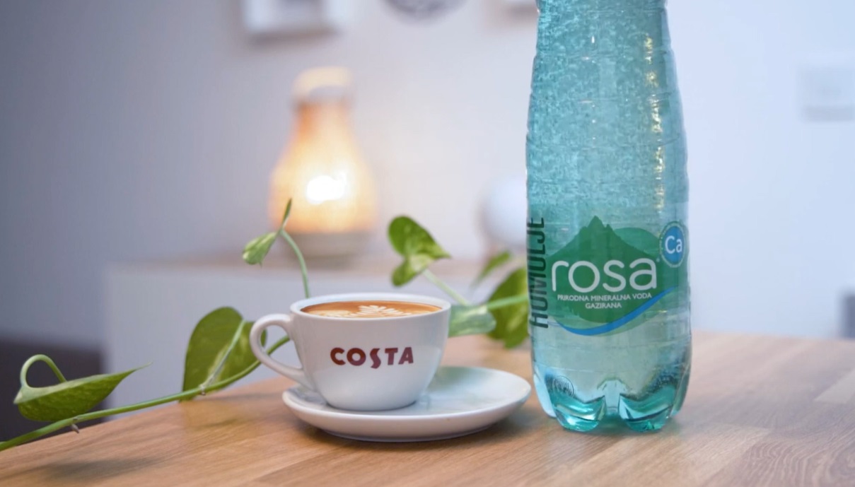 Šolja kafe Costa na tanjiriću pored flaše Rosa prirodne mineralne gazirane vode, postavljene na drvenom stolu sa dekorativnom biljkom u pozadini.