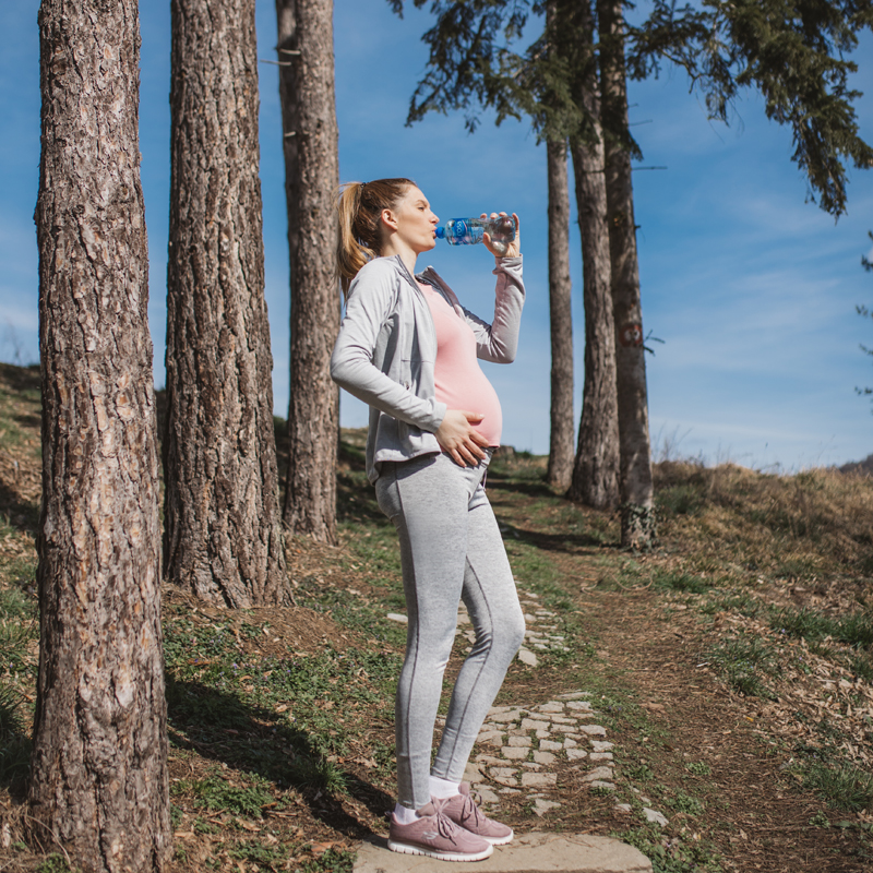 Trudnica u sportskoj odeći stoji na šumskoj stazi i pije vodu iz plastične boce