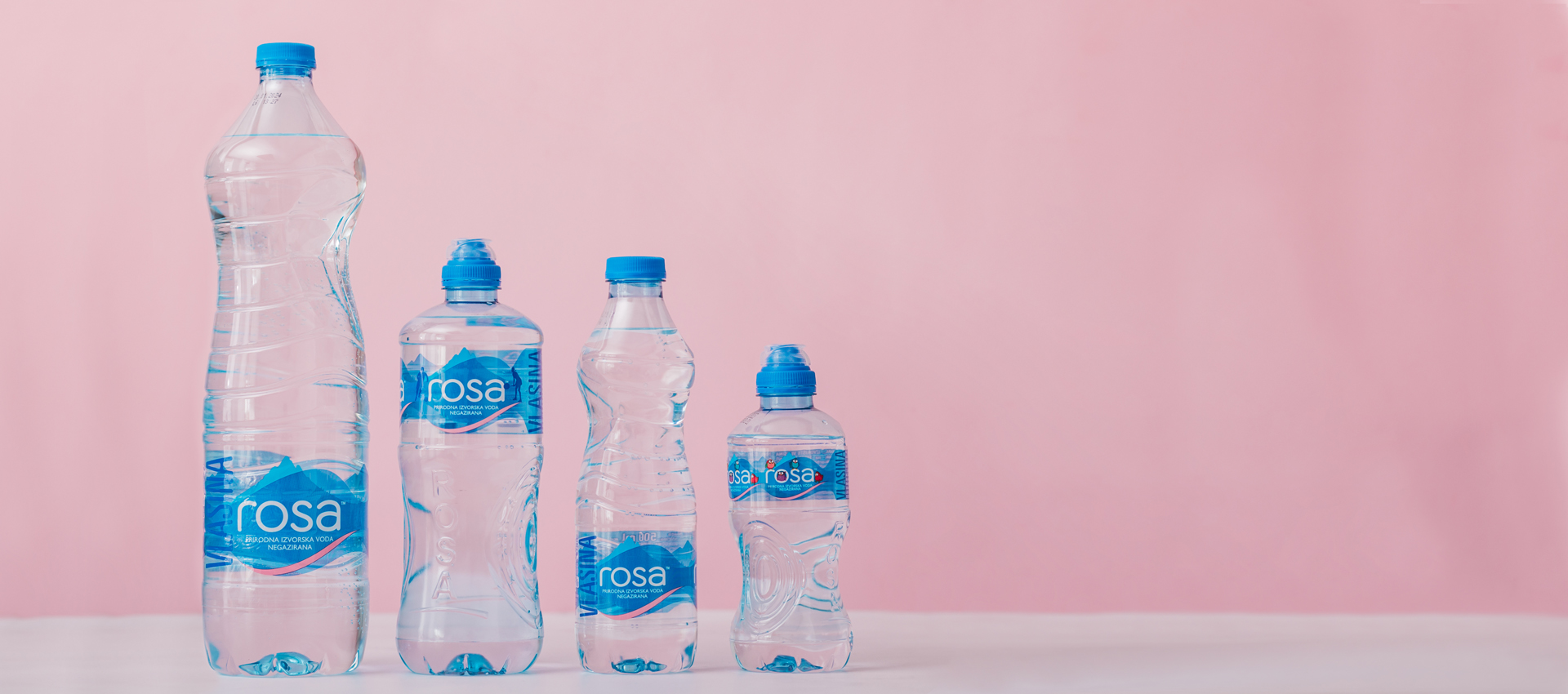 Četiri flaše Rosa vode različitih veličina poređane po visini ispred ružičaste pozadine