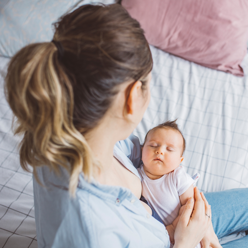 Osoba sedi na krevetu i drži bebu obučenu u belu odeću
