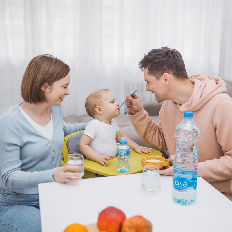 Porodična scena za stolom, odrasli hrane bebu dok na stolu stoje flaše vode, čaše i voće