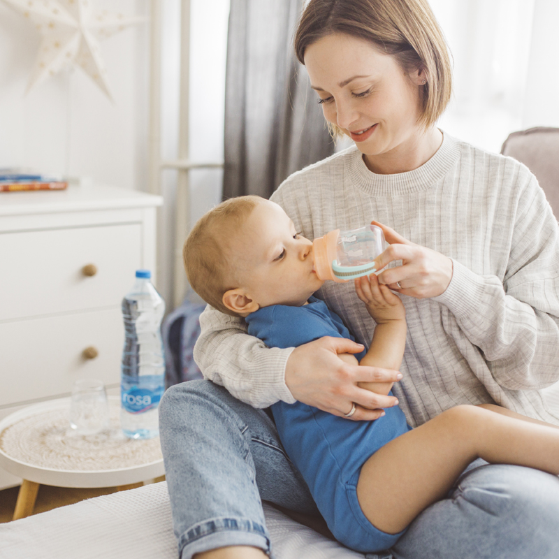 Osoba sedi na krevetu i drži dete dok mu pruža čašu vode, sa bocom Rosa vode na stočiću u pozadini