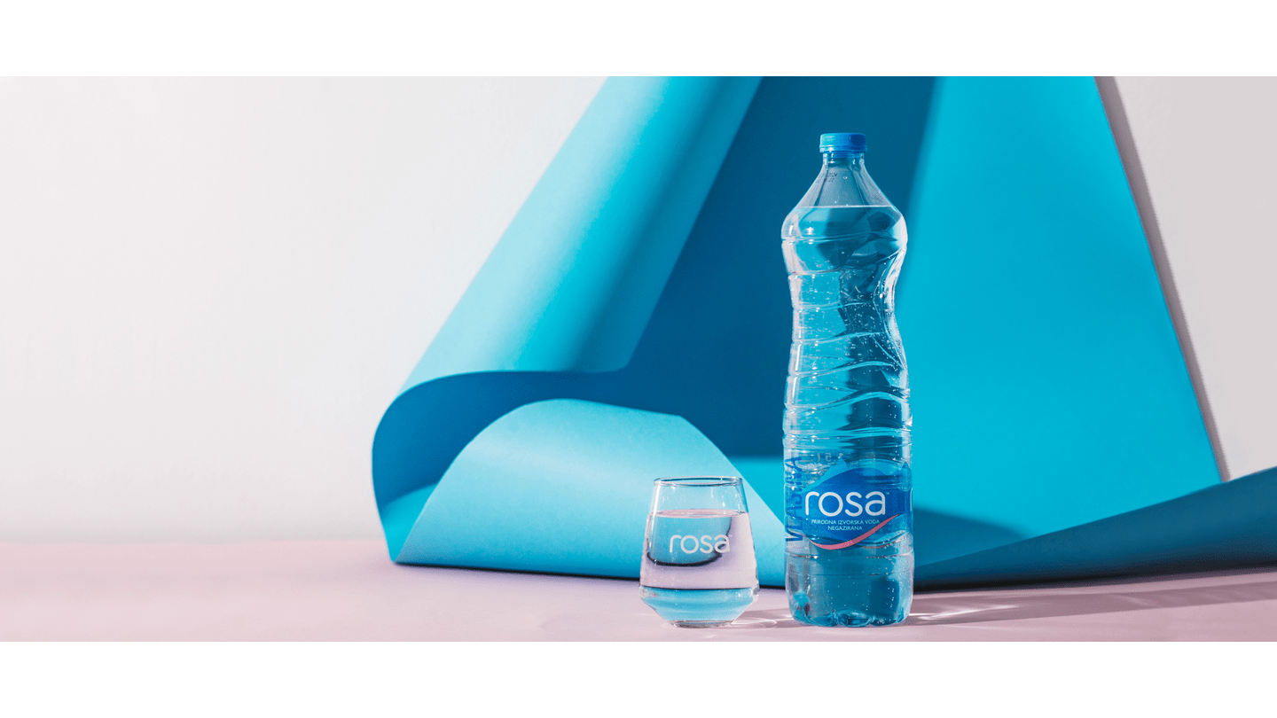 Flaša Rosa prirodne izvorske vode i čaša sa vodom postavljene ispred savijenog plavog papira na svetloj podlozi