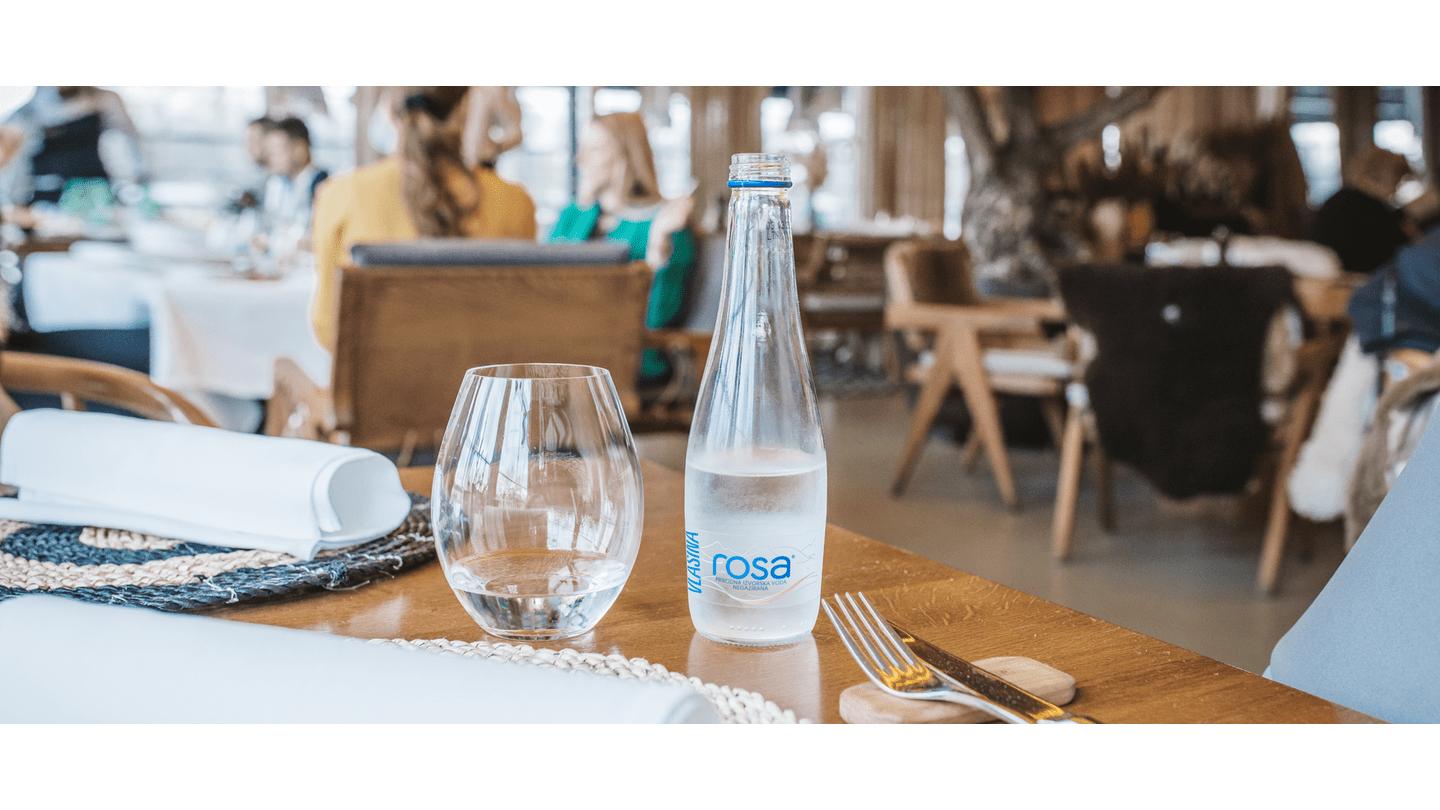 Flaša Rosa vode i čaša na postavljenom restoranskom stolu, sa zamagljenom pozadinom u kojoj se vidi ambijent restorana.