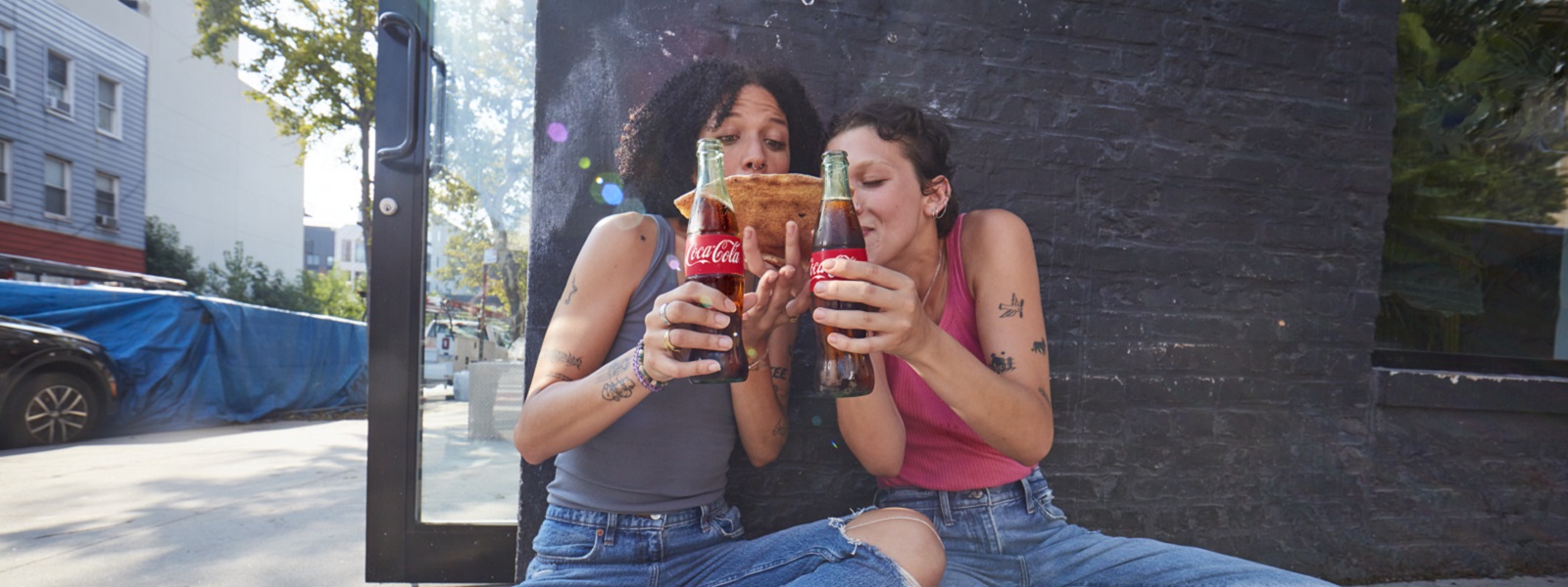 Dve osebi sedita pred temno steno, v rokah držita steklenici Coca-Cole in skupaj gledata kos pice, ki si ga delita, v sproščenem in prijateljskem trenutku..