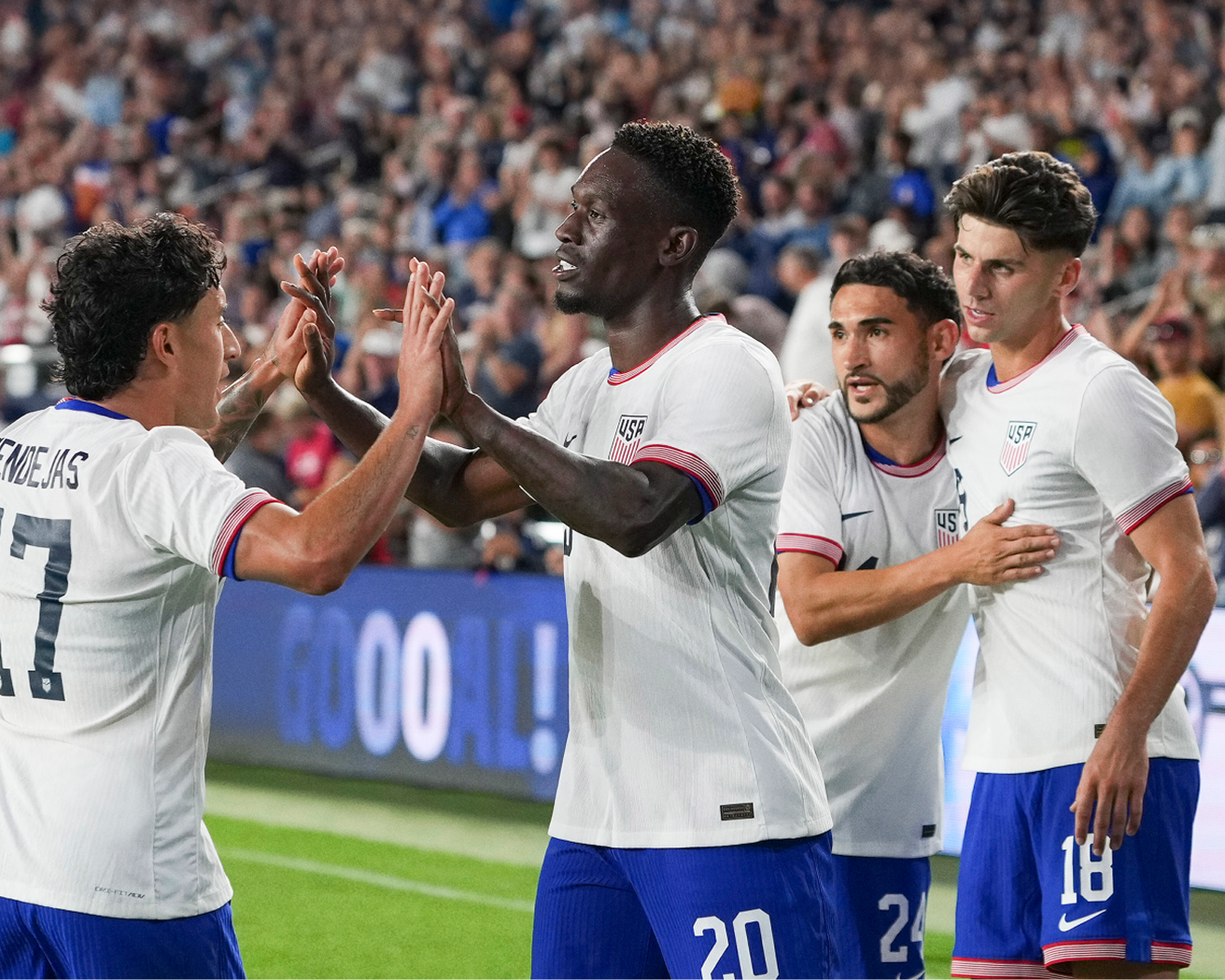 US soccer players in white jerseys and blue shorts celebrating a goal on the field, high‑fiving teammates in a packed stadium.