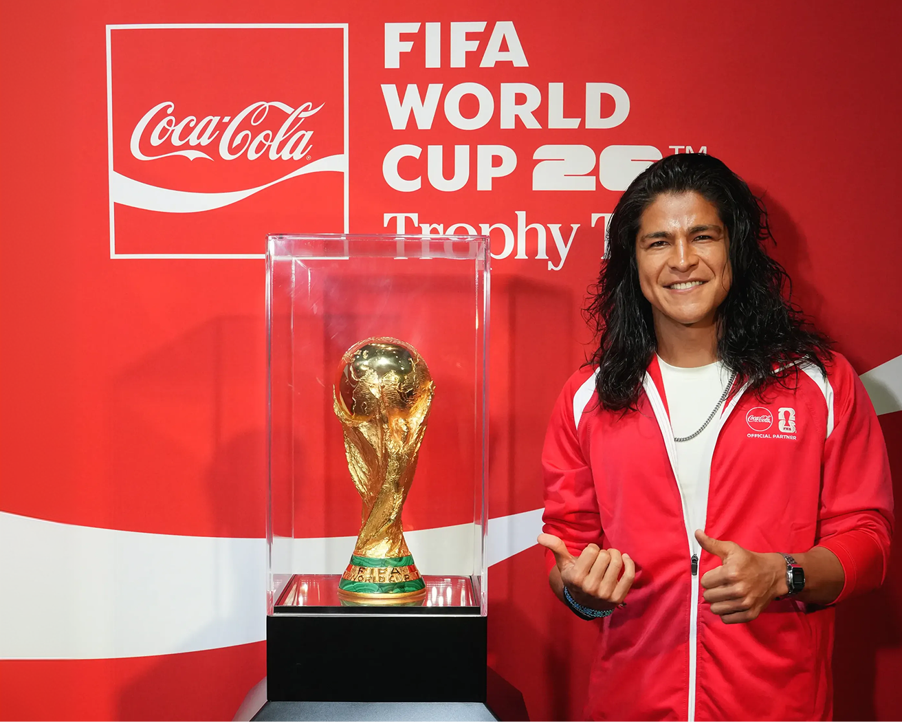 Fan in Coca-Cola jacket posing by the FIFA World Cup trophy display at U.S. Trophy Tour event