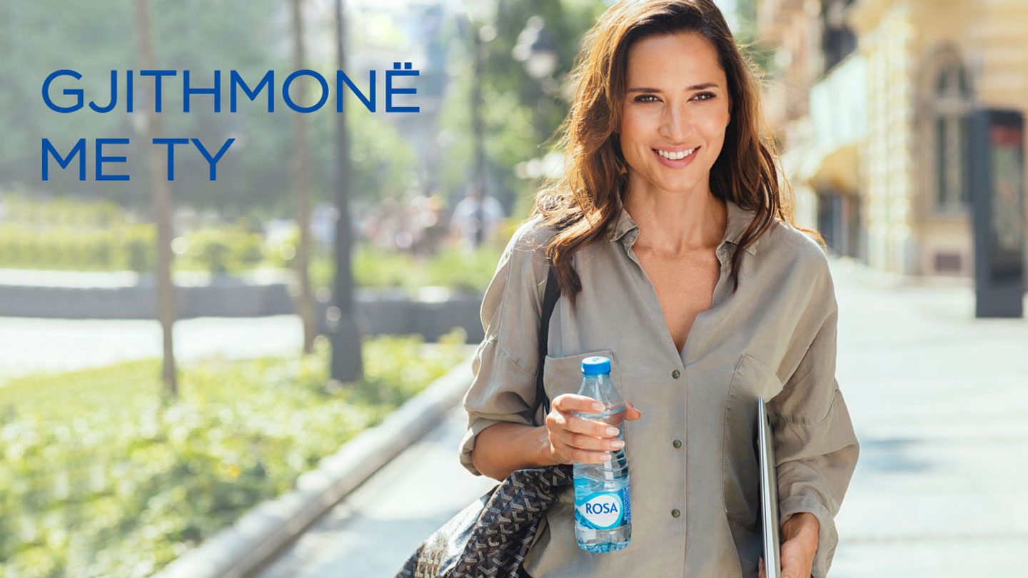 Person duke mbajtur një shishe uji Rosa dhe një laptop, me tekstin “Gjithmonë me ty” në sfond.