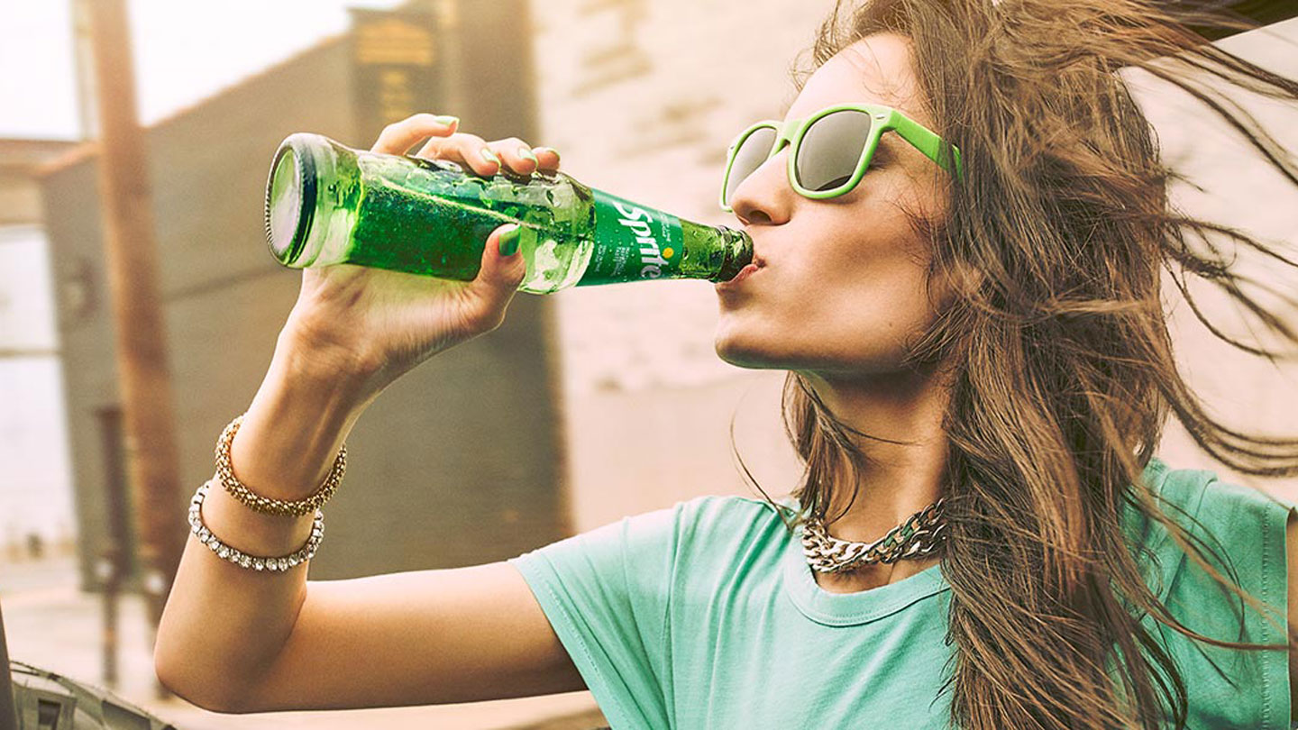 Devojka u zelenoj majici i zelenim naocarima pije Sprite iz flašice